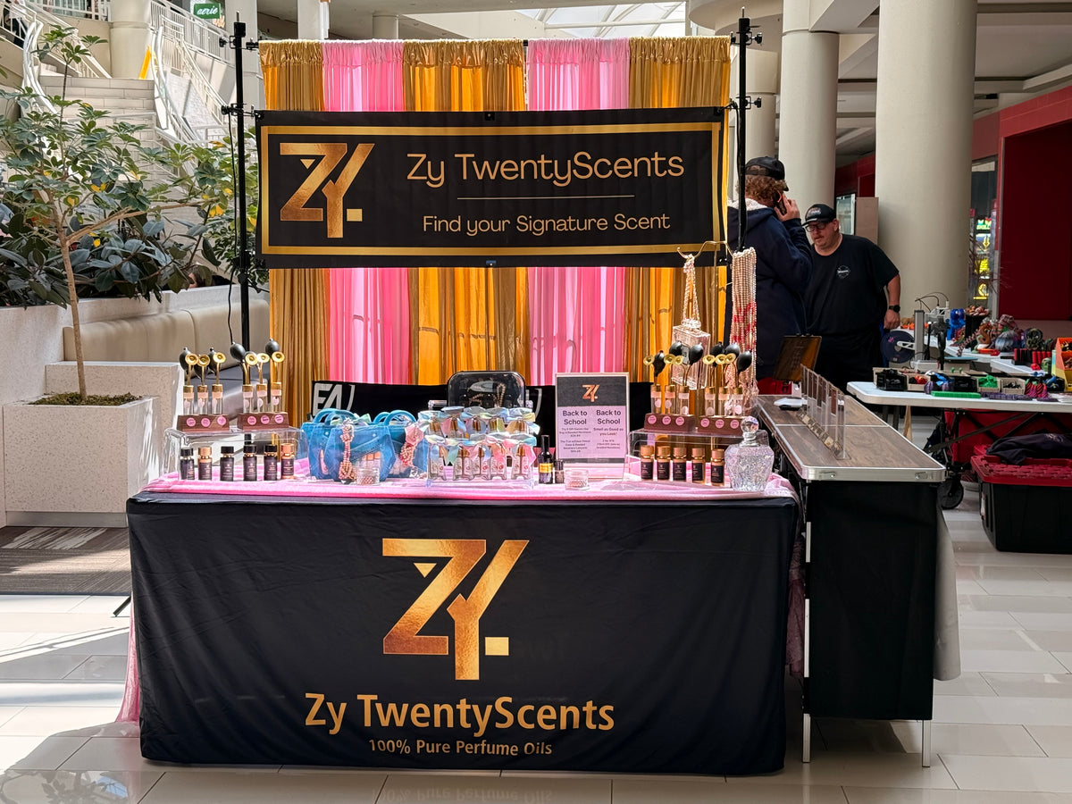 A branded Zy TwentyScents perfume display booth in a mall or event space, with a black table covered in a black-and-gold branded drape, a large hanging sign, rows of perfume bottles, and two people standing behind the counter.